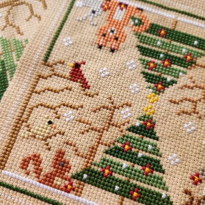 Puede incluir: Un patr&oacute;n de punto de cruz con un &aacute;rbol de Navidad verde con adornos rojos y amarillos, un cardenal, una ardilla, un zorro y un p&aacute;jaro. El patr&oacute;n est&aacute; cosido en una tela beige.