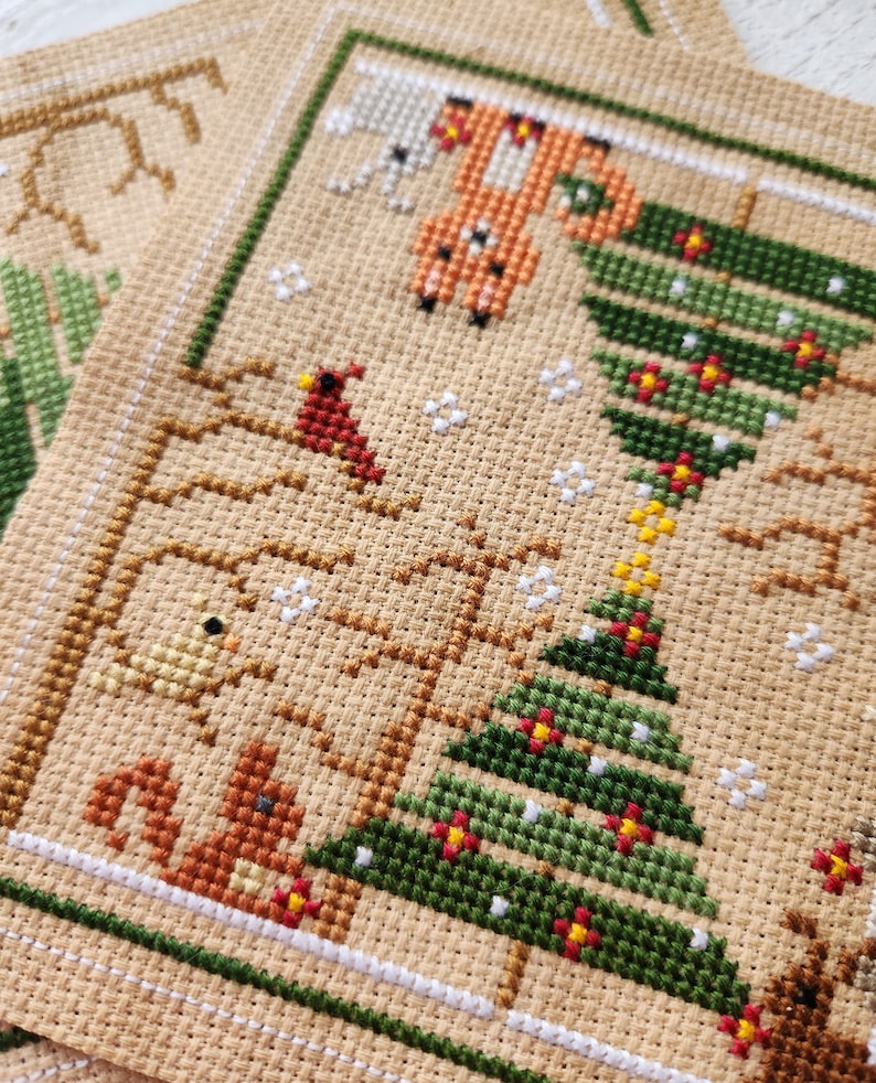 Puede incluir: Un patr&oacute;n de punto de cruz con un &aacute;rbol de Navidad verde con adornos rojos y amarillos, un cardenal, una ardilla, un zorro y un p&aacute;jaro. El patr&oacute;n est&aacute; cosido en una tela beige.
