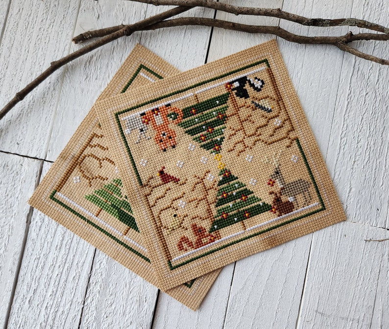Puede incluir: Dos patrones de punto de cruz beige con un tema de animales del bosque en verde, marr&oacute;n y rojo. Los patrones representan un &aacute;rbol de Navidad con adornos y animales como un zorro, una ardilla, un ciervo y un conejo.