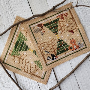 Puede incluir: Dos patrones de punto de cruz que representan escenas de invierno con &aacute;rboles, animales y copos de nieve. Los patrones est&aacute;n sobre una tela beige con hilo verde, marr&oacute;n, blanco y naranja.