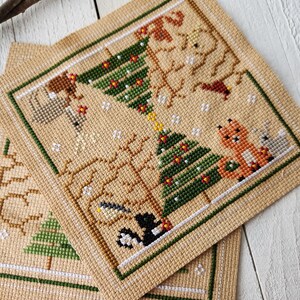 Puede incluir: Dos patrones de punto de cruz que representan escenas de invierno con &aacute;rboles, animales y copos de nieve. Los patrones est&aacute;n bordados en tela Aida beige con hilo verde, rojo, marr&oacute;n y blanco.