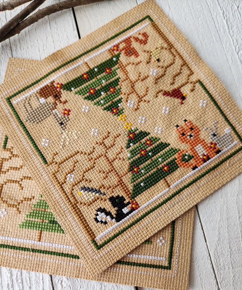 Puede incluir: Dos patrones de punto de cruz que representan escenas de invierno con &aacute;rboles, animales y copos de nieve. Los patrones est&aacute;n bordados en tela Aida beige con hilo verde, rojo, marr&oacute;n y blanco.