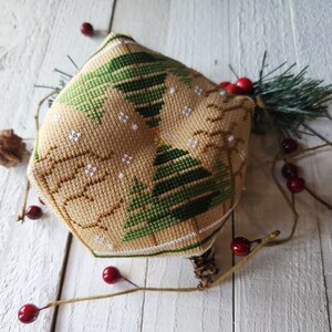 Puede incluir: Un adorno de punto de cruz beige y marr&oacute;n con un dise&ntilde;o de &aacute;rbol de Navidad verde. El adorno est&aacute; rodeado de bayas rojas y una rama de pino.