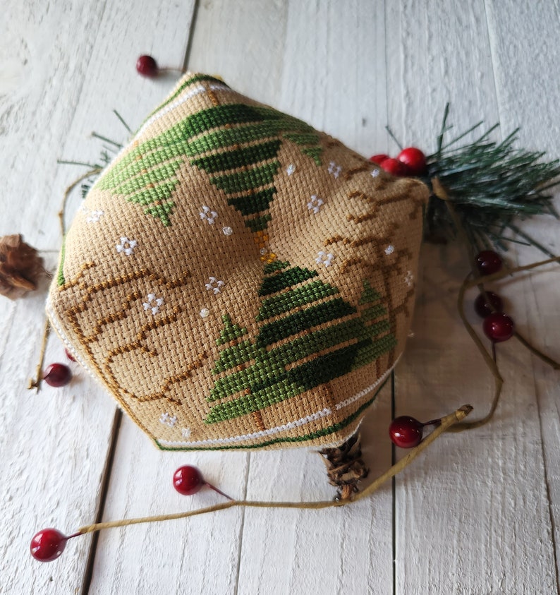 Puede incluir: Un adorno de punto de cruz beige y marr&oacute;n con un dise&ntilde;o de &aacute;rbol de Navidad verde. El adorno est&aacute; rodeado de bayas rojas y una rama de pino.