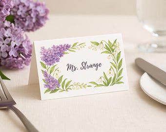 May include: A white place card with a floral border of purple and white flowers. The text "Ms. Strange" is printed in black on the card.