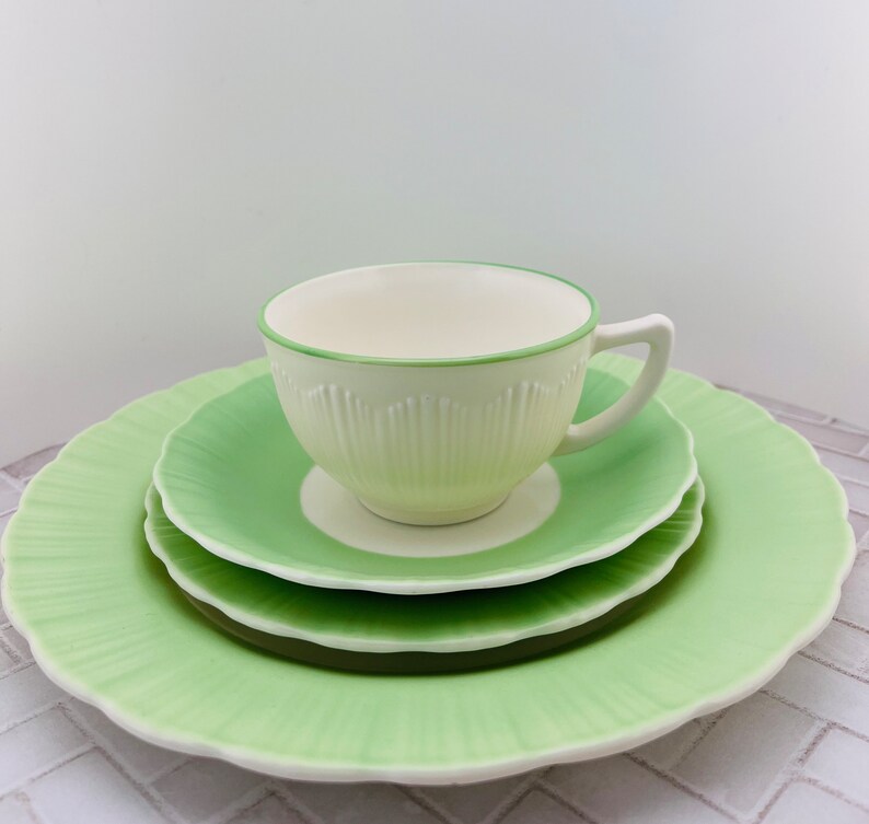 May include: A set of three green and white ceramic plates with a scalloped edge. The plates are stacked with a white teacup with a green rim on top.