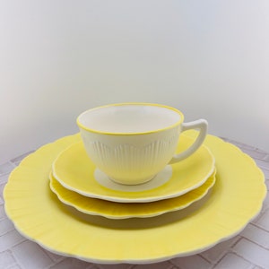 May include: A white teacup with a yellow rim sits on a yellow saucer, which is on a larger yellow saucer. All three pieces have a scalloped edge.