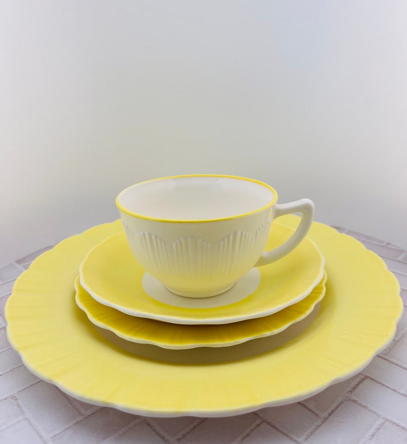 May include: A white teacup with a yellow rim sits on a yellow saucer, which is on a larger yellow saucer. All three pieces have a scalloped edge.