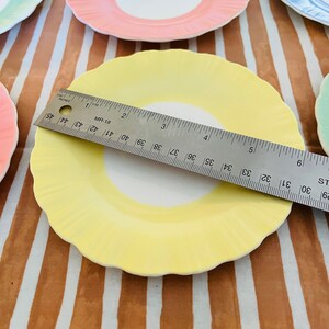 May include: A set of four pastel-colored ceramic plates with scalloped edges. The plates are yellow, pink, blue, and green. A ruler is placed on top of the yellow plate.