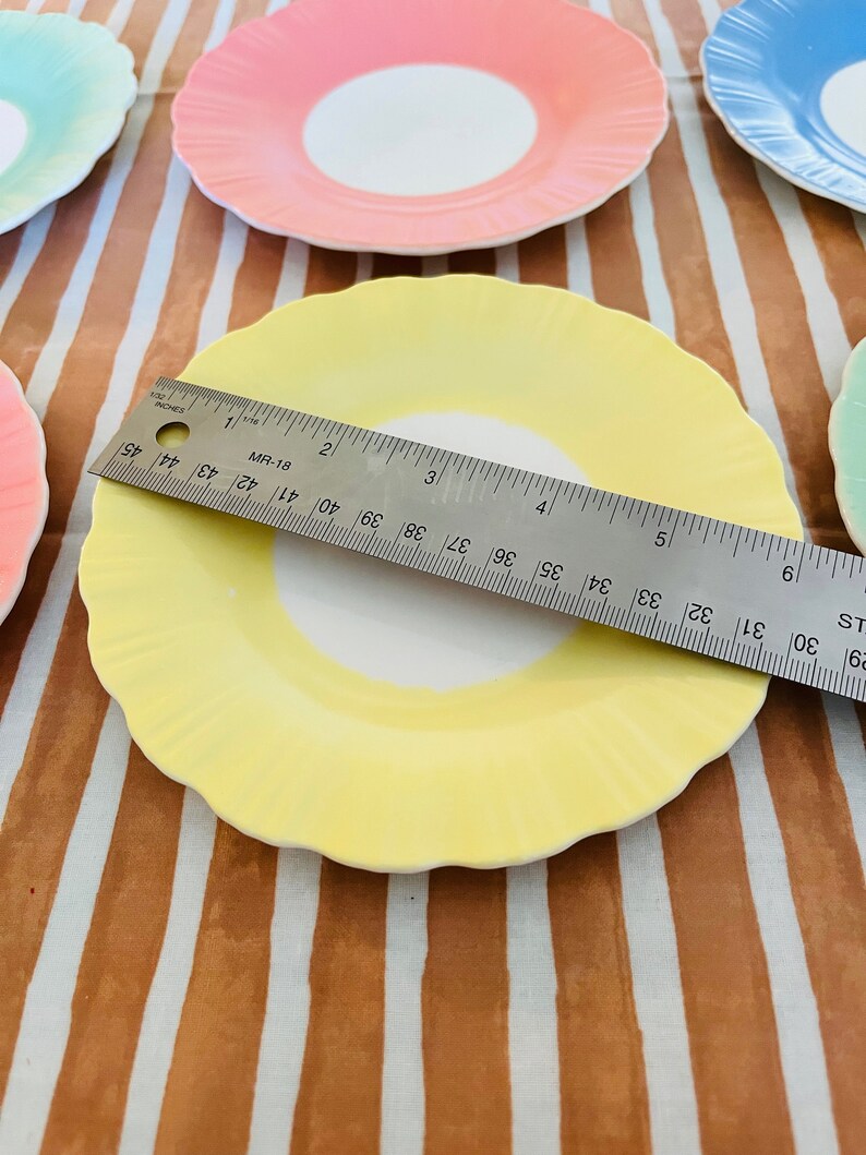 May include: A set of four pastel-colored ceramic plates with scalloped edges. The plates are yellow, pink, blue, and green. A ruler is placed on top of the yellow plate.
