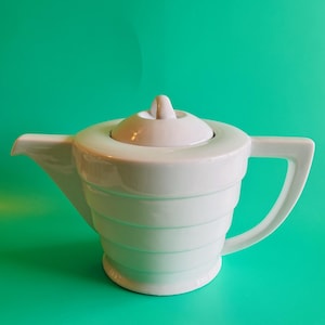 May include: A white ceramic teapot with a lid and handle. The teapot has a unique design with horizontal ridges around the body. The spout is on the left side, and the handle is on the right. The background is a solid teal color.