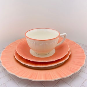 May include: A set of three peach-colored ceramic plates with a scalloped edge. The plates are stacked with a white teacup with a peach-colored rim on top.
