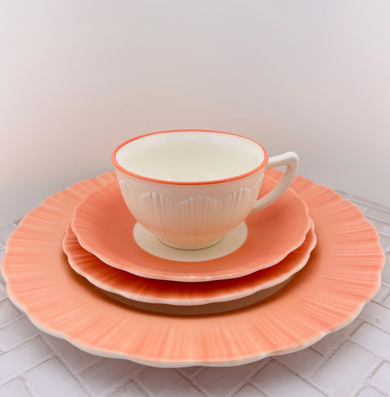 May include: A set of three peach-colored ceramic plates with a scalloped edge. The plates are stacked with a white teacup with a peach-colored rim on top.