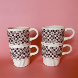 May include: Four white ceramic mugs stacked in two pairs. Each mug features a dark brown checkered pattern with small white flower icons inside every other square. The base of each mug has a vertical ribbed texture around the bottom section.
