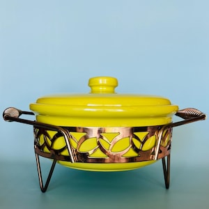 May include: A bright yellow ceramic casserole dish with a lid, set in a decorative copper-toned metal stand. The stand features a leaf-like cutout design. The handles are also copper-toned.