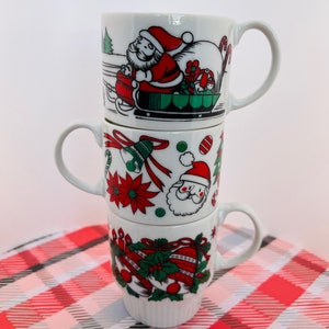 May include: A set of three white ceramic mugs with Christmas-themed designs. The mugs feature Santa Claus, bells, holly, and Christmas trees in red, green, and white.