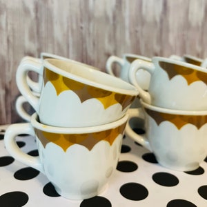 May include: A stack of six white ceramic coffee cups with a gold and brown scalloped design. The cups are stacked on a black and white polka dot surface.