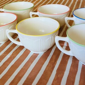 May include: Set of seven white ceramic teacups with colored rims. The rims are green, yellow, blue, and pink. The cups are on a striped brown and white surface.