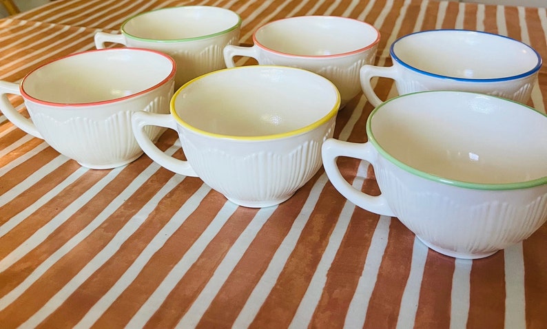 May include: Set of seven white ceramic teacups with colored rims. The rims are green, yellow, blue, and pink. The cups are on a striped brown and white surface.