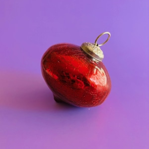 May include: A red, heart-shaped Christmas ornament with a silver cap and loop for hanging. The ornament has a textured, crackled surface, and is set against a purple background. A festive holiday decoration.
