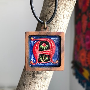Puede incluir: Un colgante de madera con una letra A roja pintada a mano sobre un fondo azul. La letra está decorada con detalles dorados y una pequeña flor verde.