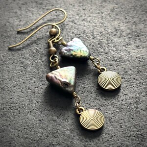 May include: A pair of handcrafted earrings featuring iridescent, irregularly shaped beads and small, round, spiral-patterned charms. The earrings are suspended from gold-colored ear wires and connected with delicate chains.
