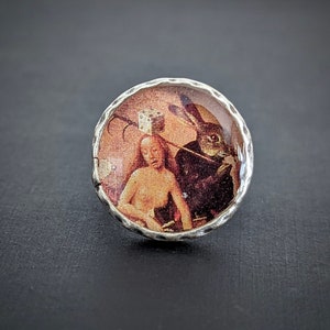 May include: A silver ring with a glass cabochon featuring a vintage illustration of a person with a rabbit. The illustration is in shades of brown, red, and white.