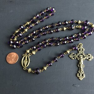 Puede incluir: Un rosario de cristal morado con detalles dorados y un crucifijo dorado con una figura de Jesucristo.