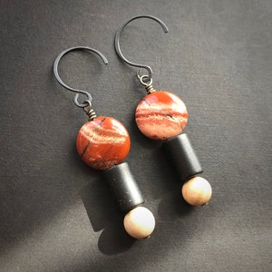 May include: Handmade earrings with a red and cream-coloured circular stone, a black cylindrical bead, and a small cream-coloured bead. The earrings hang from a dark metal hook. The design is minimalist and modern.