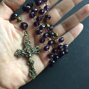 Puede incluir: Un rosario de cristal morado con un crucifijo de bronce. El crucifijo presenta un dise&ntilde;o detallado y una figura de Jesucristo.