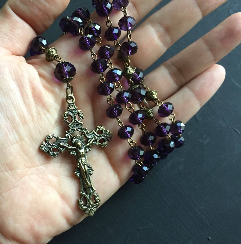 Puede incluir: Un rosario de cristal morado con un crucifijo de bronce. El crucifijo presenta un dise&ntilde;o detallado y una figura de Jesucristo.