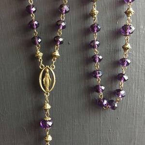 Puede incluir: Un collar de rosario morado y dorado con un peque&ntilde;o colgante ovalado que representa a la Virgen Mar&iacute;a.
