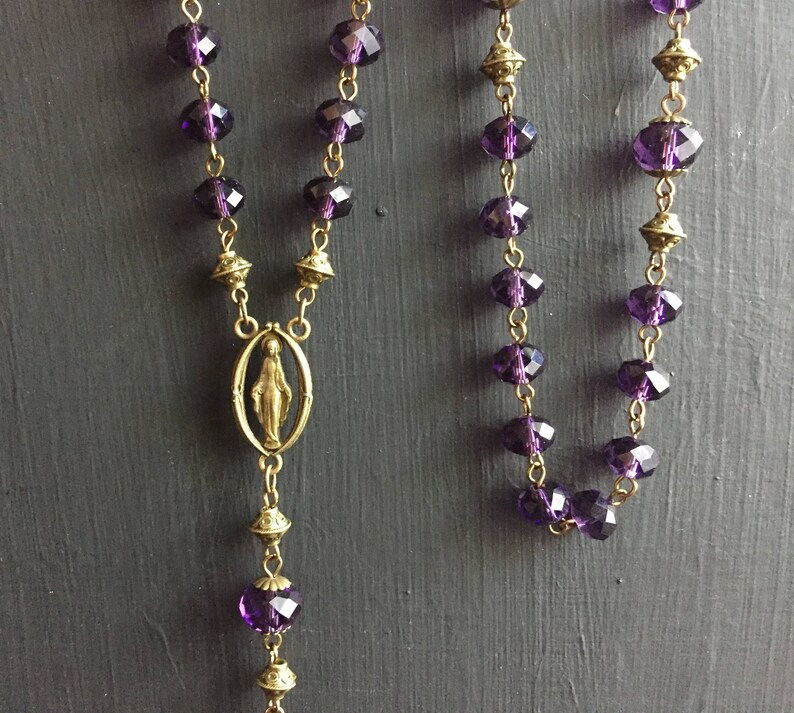 Puede incluir: Un collar de rosario morado y dorado con un peque&ntilde;o colgante ovalado que representa a la Virgen Mar&iacute;a.