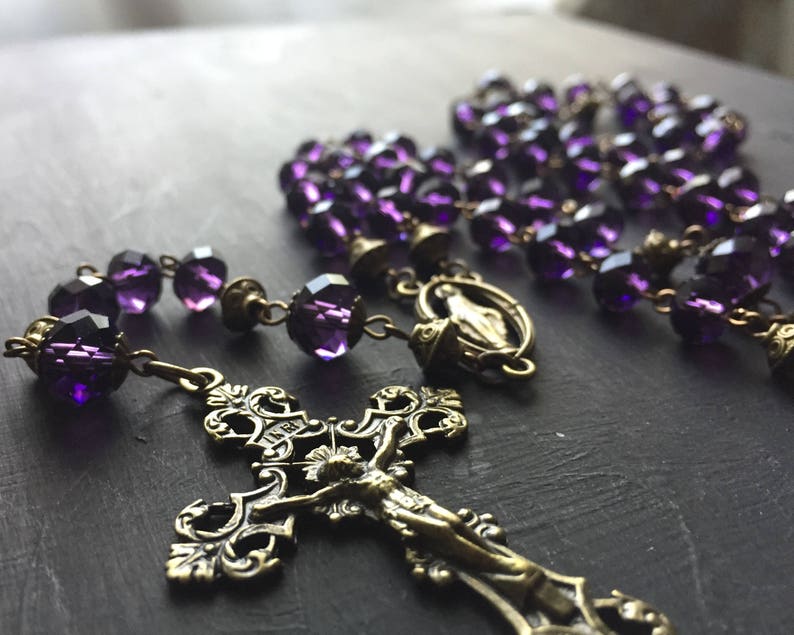 Puede incluir: Un rosario de cristal morado con un crucifijo de lat&oacute;n y un amuleto de lat&oacute;n de la Virgen Mar&iacute;a. Las cuentas del rosario son facetadas y tienen un color morado oscuro.