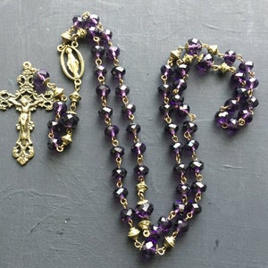 Puede incluir: Un rosario de cristal morado con un crucifijo dorado y un peque&ntilde;o dije dorado. El rosario est&aacute; hecho de cuentas de vidrio facetadas y tiene una cadena dorada.