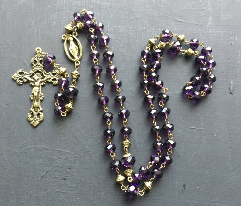 Puede incluir: Un rosario de cristal morado con un crucifijo dorado y un peque&ntilde;o dije dorado. El rosario est&aacute; hecho de cuentas de vidrio facetadas y tiene una cadena dorada.