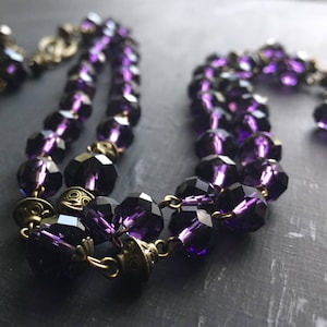 Puede incluir: Un collar de doble cadena hecho de cuentas de vidrio facetado de color morado oscuro y detalles met&aacute;licos de oro antiguo.