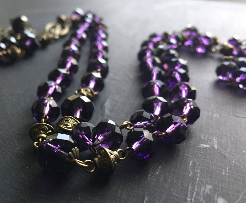 Puede incluir: Un collar de doble cadena hecho de cuentas de vidrio facetado de color morado oscuro y detalles met&aacute;licos de oro antiguo.