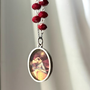 May include: A silver pendant necklace with an oval pendant displaying a small image of a figure. The necklace features a chain of faceted, deep red beads. The pendant is suspended from a silver chain.