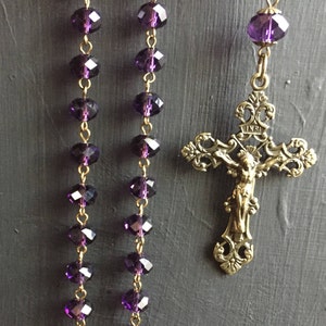 Puede incluir: Una cadena de oro con un crucifijo y cuentas de vidrio morado. El crucifijo tiene un dise&ntilde;o detallado y la inscripci&oacute;n "INRI" en la parte superior.