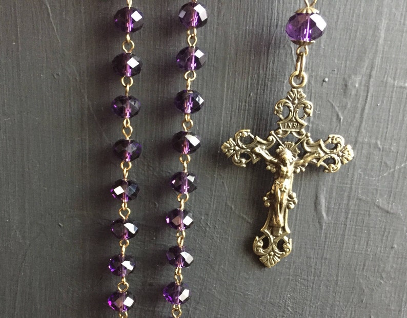 Puede incluir: Una cadena de oro con un crucifijo y cuentas de vidrio morado. El crucifijo tiene un dise&ntilde;o detallado y la inscripci&oacute;n "INRI" en la parte superior.