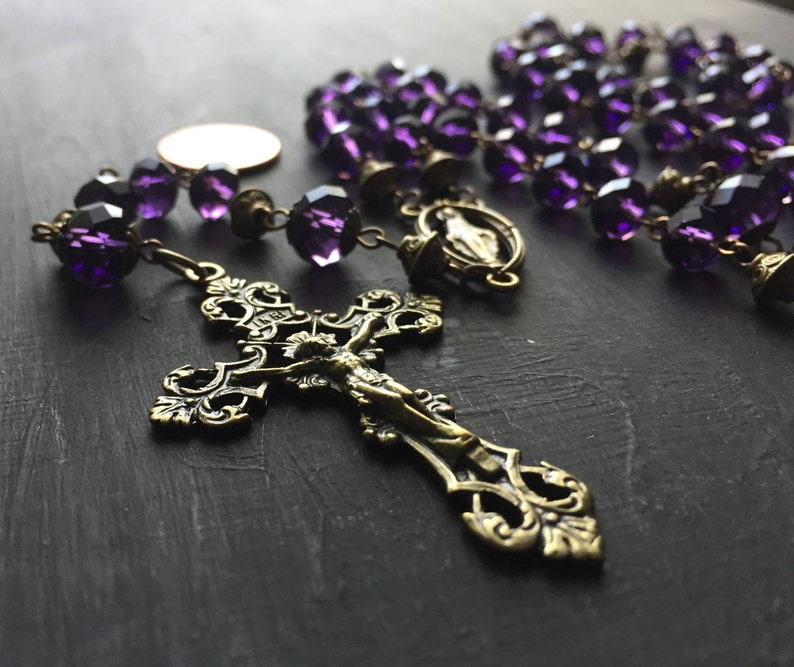 Puede incluir: Un rosario de cristal morado con un crucifijo de lat&oacute;n y un dije de lat&oacute;n de la Virgen Mar&iacute;a. Las cuentas del rosario son facetadas y tienen un color morado oscuro.