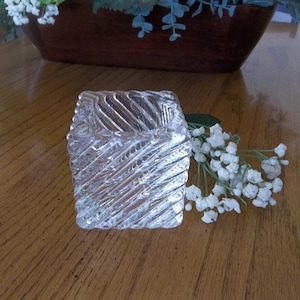 May include: A clear, square glass ashtray with a textured, diagonal pattern. The ashtray sits on a wooden surface, with a floral arrangement and white flowers in the background.