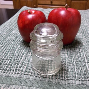 May include: A clear glass bottle stopper with three tiers. The stopper is sitting on a green and white patterned towel with two red apples behind it.