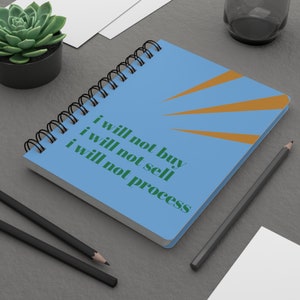 May include: A blue spiral notebook with a sunburst design and the text "i will not buy i will not sell i will not process" in green.