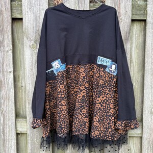 May include: A black long-sleeve top featuring a brown and black animal print lower section, a black polka-dot mesh ruffle at the hem, and two denim patches on the chest. One patch displays the word love in white script lettering.
