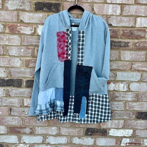 May include: A gray hooded sweatshirt with patchwork details. The patches include red floral, black and white plaid, and denim fabrics. The bottom hem features a black and white checkered pattern and a blue ruffled fabric.