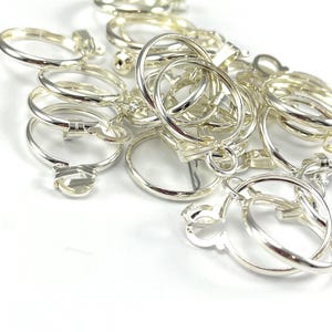 Peut inclure: Un ensemble de bagues en métal argenté brillant, certaines avec des formes décoratives. Les bagues sont éparpillées sur une surface blanche, reflétant la lumière. Les bagues ont un mécanisme de fermeture, suggérant qu'elles sont pour la bijouterie ou l'artisanat.