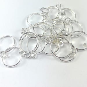 以下が含まれることがあります： 小さなループと留め金が付いた、銀色の金属製の丸いチャームのコレクション。チャームは白い表面に散らばっており、一部は重なっています。チャームは反射性があり、新品のようです。