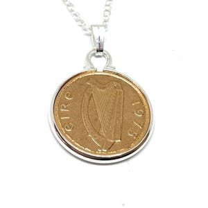 May include: A gold-colored Irish coin pendant necklace with a silver chain and a silver border around the coin. The coin features a harp and the year 1975.